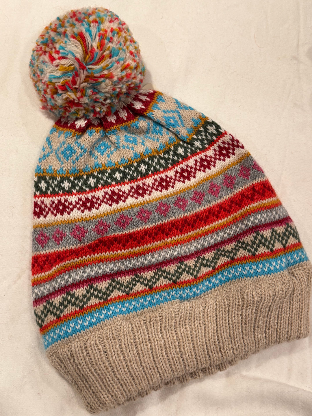 Alpine Confetti hand-knit beanie with narrow Fair Isle bands in turquoise, crimson, gold and sage on an oatmeal crown, topped with a multicolour flecked pom