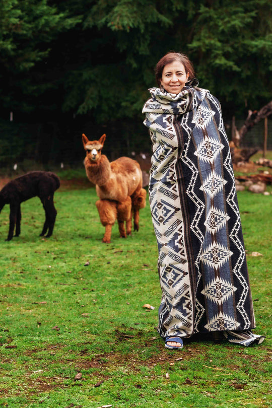 Woman wrapped in the Cuzco Stone Alpaca Wool Blanket with alpacas grazing in the background at Enchanted Farms.