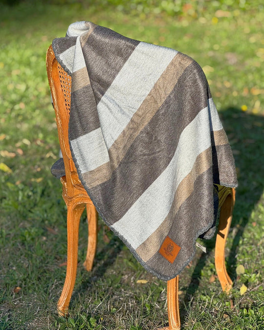 Driftwood brushed alpaca throw blanket in charcoal, silver grey and taupe stripes, draped over a cane chair at Enchanted Farms