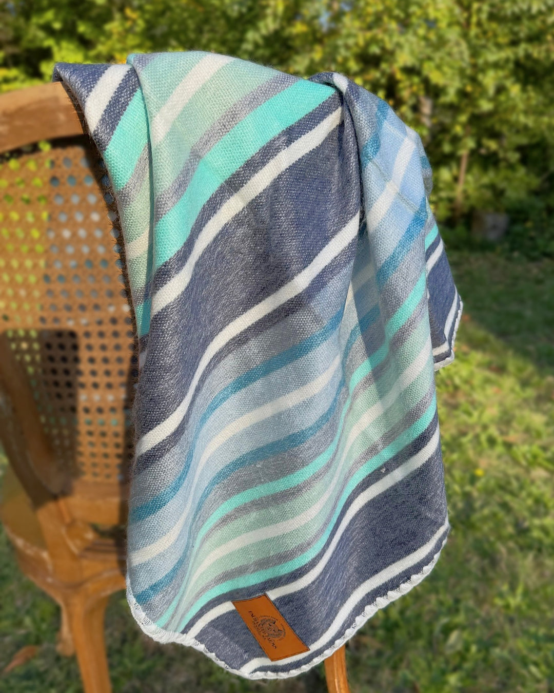 Seaglass brushed alpaca throw blanket in seafoam, sky blue, grey and navy stripes, draped over a cane chair at Enchanted Farms